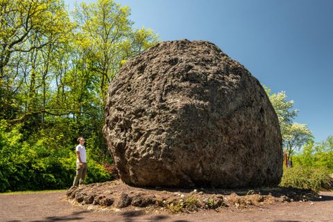 Ein großer Felsen steht in einer grünen Umgebung. Eine Person betrachtet den Felsen bei sonnigem Wetter.