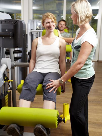 Eine Frau trainiert an einem Fitnessgerät und lächelt. Eine Trainerin gibt ihr Anweisungen und Unterstützung.