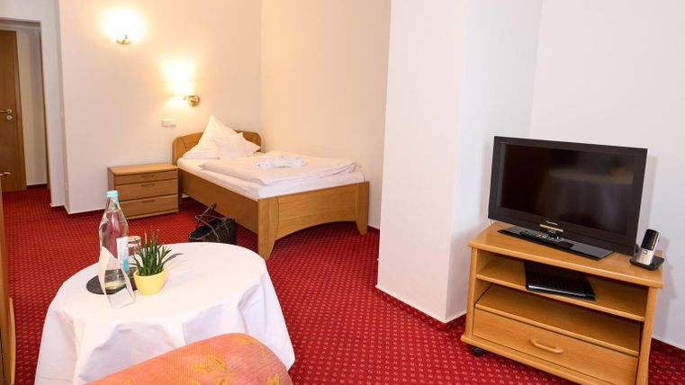 Ein einfaches Hotelzimmer mit einem Einzelbett, einem Fernseher und einem kleinen Tisch. Der Teppich ist rot und die Wände sind hell gestaltet.