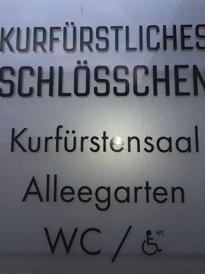 Ein Schild mit dem Text "Kurfürstliches Schlösschen" und Informationen zu einem Veranstaltungsraum sowie einem WC. Es enthält auch Hinweise für die Barrierefreiheit.