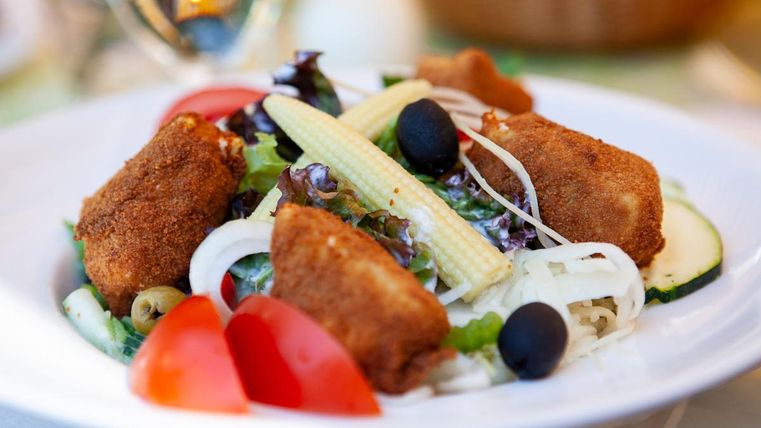 Ein Teller mit frischem Salat, knusprigen Croquettes und verschiedenen Gemüsesorten. Dazu sind Tomaten, Oliven und Baby-Mais dekorativ angerichtet.