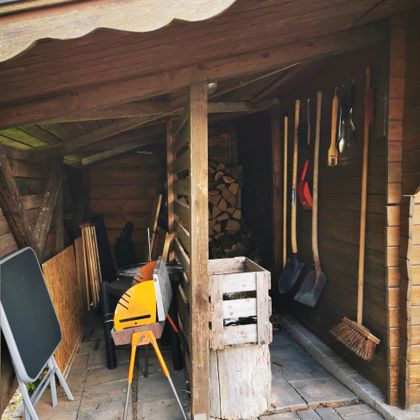 Eine kleine Gartenhütte mit Werkzeugen an der Wand und Holzstapeln. Auf dem Boden stehen verschiedene Gerätschaften und eine Holzkiste.