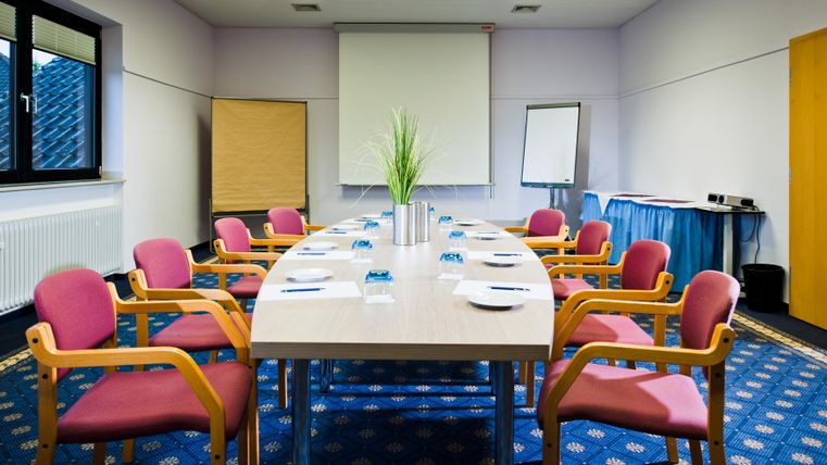 Ein moderner Besprechungsraum mit einem großen Tisch und roten Stühlen. Es gibt einen Projektor und eine Pflanzen Dekoration in der Mitte.