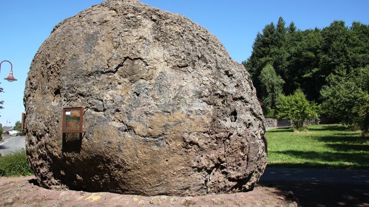 Ein großer, runder Felsen steht in einer grünen Wiese. Der Himmel ist klar und blau.