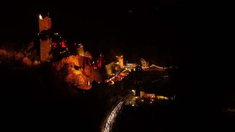Een verlichte kasteel op een heuvel in de nacht. Op de voorgrond zijn lichtsporen van voertuigen op de weg te zien.