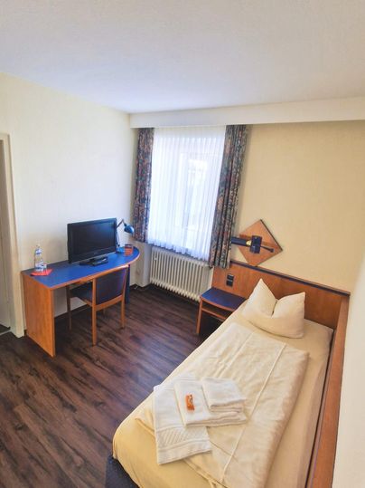 Ein einfaches Hotelzimmer mit einem Einzelbett und einem Schreibtisch. Es gibt einen Fernseher und viel natürliches Licht durch das Fenster.