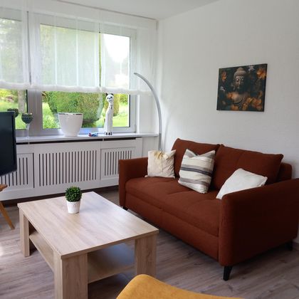 A cozy living room with a brown sofa and a wooden table. The large windows provide plenty of light and a view of the garden.