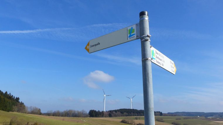 Wegweiser an der Lieserquelle mit Windrädern im Hintergrund.