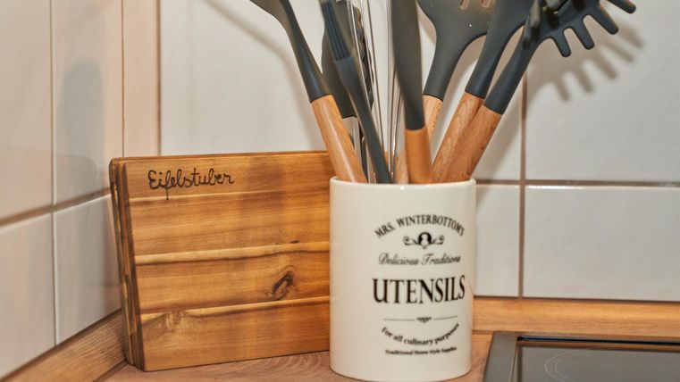 Ein Küchenschrank mit einem Töpfehalter und verschiedenen Kochutensilien. Der Behälter ist weiß mit einem Schriftzug und die Utensilien haben Holzelemente.