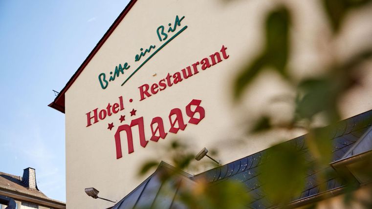 Das Bild zeigt die Außenwand eines Hotels mit der Aufschrift „Hotel · Restaurant Maas“. Der Blick ist teilweise durch Blätter eines Baumes verdeckt.