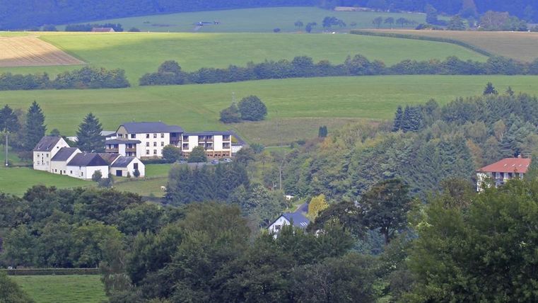 Eine malerische Landschaft mit grünen Wiesen und sanften Hügeln. Im Vordergrund sind einige Häuser und Bäume sichtbar.