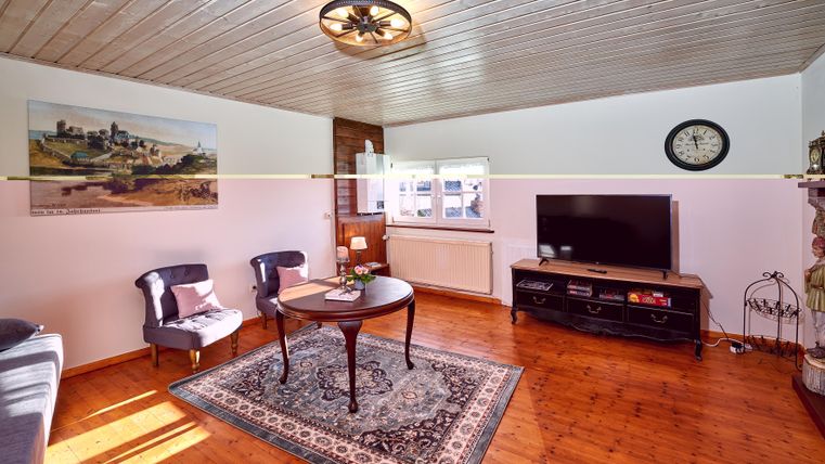 Ein gemütliches Wohnzimmer mit Holzboden und einer hellen Decke. Es gibt einen Tisch, zwei Stühle und einen Fernseher an der Wand.