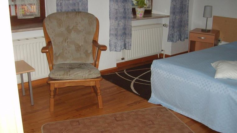Ein einfaches Zimmer mit einem Bett und einem bequemen Stuhl. Es gibt Fenster mit Vorhängen und einen kleinen Tisch.