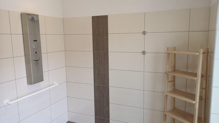 Ein heller Duschbereich mit weißen Fliesen und einem Holzregal. An der Wand sind eine Duscharmatur und Haltegriffe angebracht.
