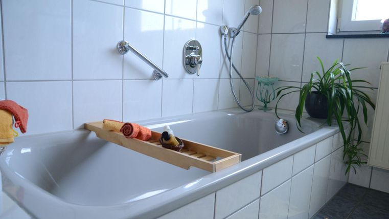 Ein helles Badezimmer mit einer Badewanne und weißen Fliesen. Auf der Badewanne liegt ein Holztablett mit Badeutensilien und eine Pflanze steht im Hintergrund.