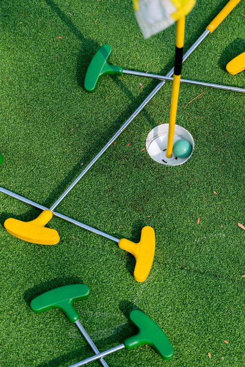 <p>In der Mitte des Bildes sieht man ein Loch, indem ein grüner Golfball liegt. Um das Loch ist grüner Kunstrasen ausgelegt, auf dem mehrere Schläger liegen. Die Schläger haben gelbe und grüne Schlägerköpfe, der Schaft ist metallfarbend.</p>