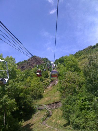 Eine Seilbahn fährt durch eine grüne Landschaft. Im Hintergrund sind Bäume und ein blauer Himmel zu sehen.