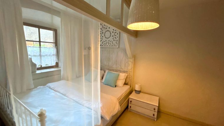 Ein gemütliches Schlafzimmer mit einem Bett, das von einem weißen Vorhang umgeben ist. Ein Nachttisch und eine warm beleuchtete Deckenlampe verleihen dem Raum eine einladende Atmosphäre.
