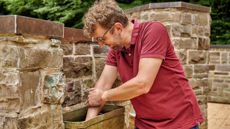 Ein Mann steht neben einer Steinmauer und wäscht seine Hände in einem Wasserbecken. Die Umgebung ist grün und naturnah.