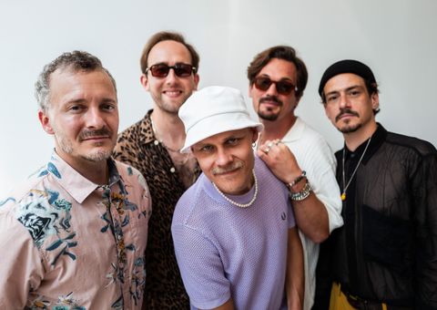 Eine Gruppe von sechs Musikern posiert in einem gemütlichen Raum. Sie stehen und sitzen um einen roten Sessel und tragen lässige Kleidung.