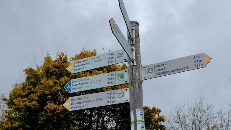 Ein Wegweiser mit verschiedenen Richtungshinweisen und Entfernungen. Im Hintergrund sind Bäume zu sehen.