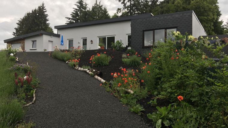 Ein modernes Haus mit einem gepflegten Garten und bunten Blumen. Der Weg zum Haus ist mit Kies bedeckt und von Bäumen umgeben.