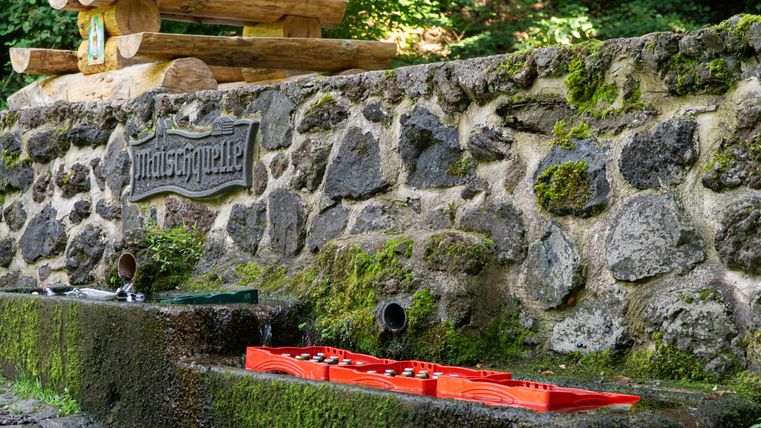 Eine Mauer, an der ein Schild mit der Inschrift "Maischquelle" hängt. Auf der Mauer steht ein Holztisch mit zwei Bänken. Unter der Mauer liegen leere Glasflaschen und Getränkekisten.