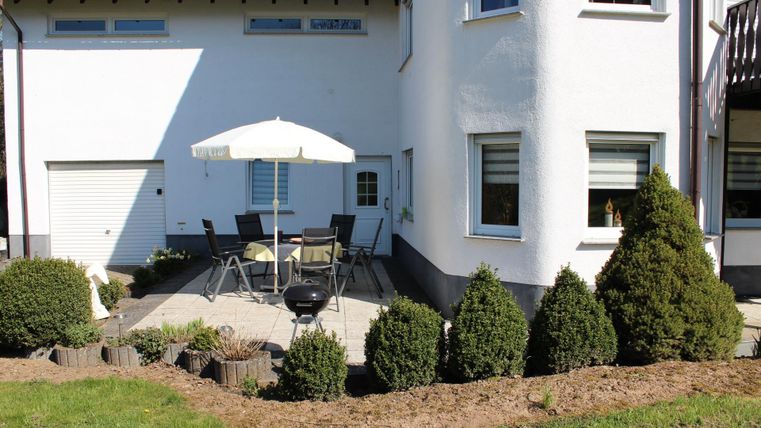 Ein gemütlicher Terrassenbereich mit einem Tisch und Stühlen unter einem Sonnenschirm. Einige kleine Bäume und ein Grill sind ebenfalls sichtbar.