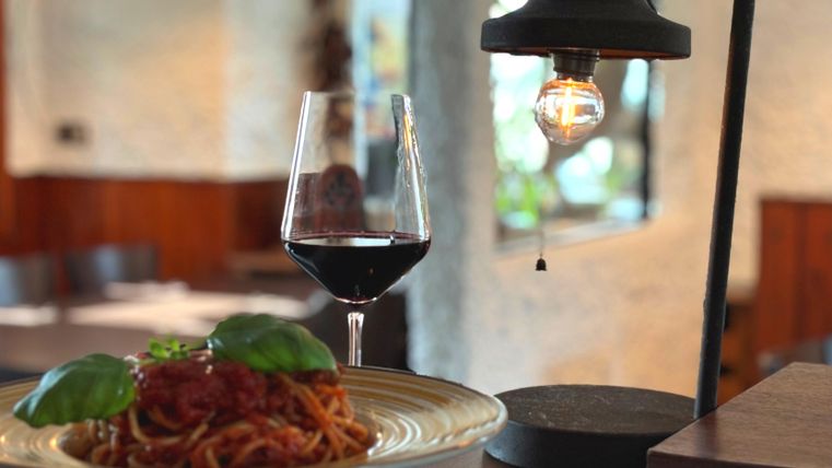 Ein Teller Spaghetti mit Tomatensauce und frischem Basilikum. Daneben steht ein Glas Rotwein unter einer hängenden Lampe.