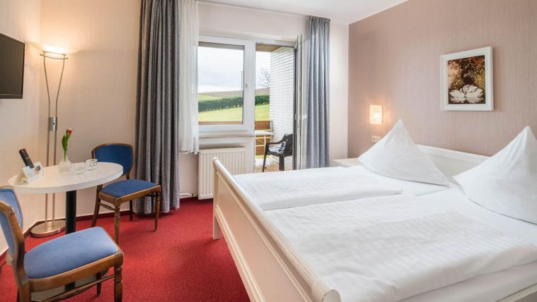 Ein gemütliches Zimmer mit einem Doppelbett und zwei Kissen. Am Tisch stehen ein paar Stühle und durch das Fenster sieht man eine grüne Landschaft.