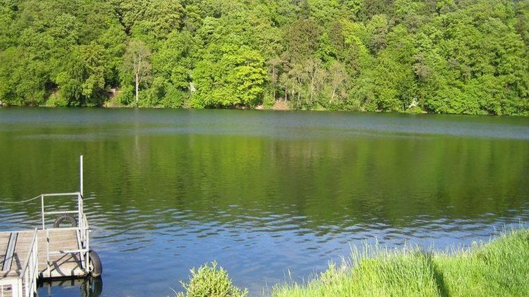 Ein ruhiger See umgeben von üppigem Grün und Bäumen. Ein kleiner Steg führt direkt zum Wasser.