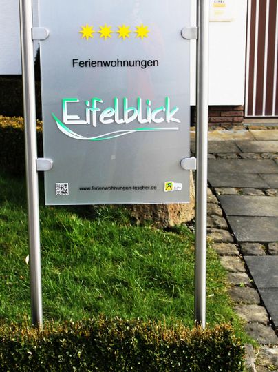 Ein Schild mit der Aufschrift "Ferienwohnungen Eifelblick". Es zeigt drei Sterne und ist in einer grünen Umgebung aufgestellt.