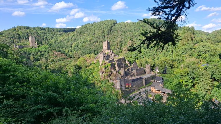 Eine alte Burgruine eingebettet in üppiges grünes Land. Im Hintergrund sind Hügel und ein klarer blauer Himmel zu sehen.