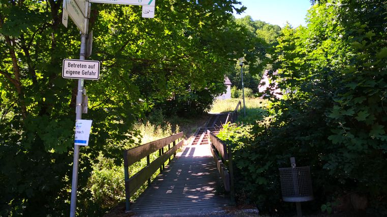 Ein schmaler Holzsteg führt durch einen grünen, bewaldeten Bereich. Schilder weisen auf das Zentrum und den Bahnhof hin.