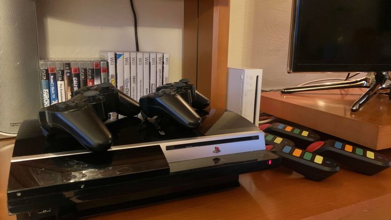 Eine Spielekonsole mit zwei Controllern steht auf einem Regal. Daneben sind Spiel-CDs und eine Fernbedienung zu sehen.