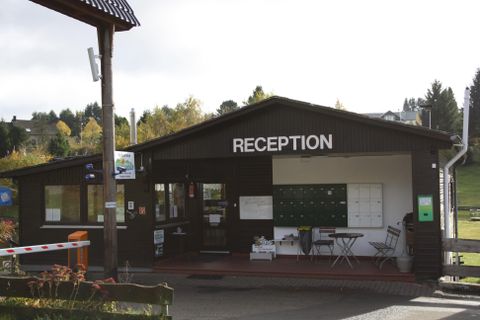 Eine Empfangshalle aus Holz mit dem Schriftzug „RECEPTION“. Vor dem Gebäude stehen Tische und Stühle.