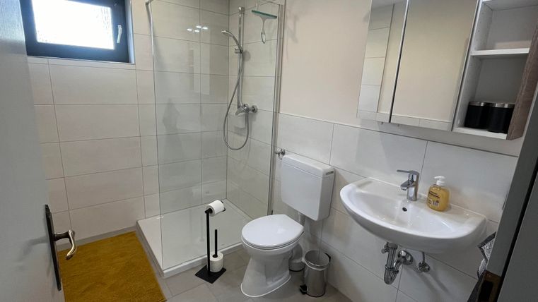 Ein modernes Badezimmer mit einer Duschkabine und einem Waschbecken. Die Wände sind hell gefliest, und es gibt eine kleine Fußmatte in einem warmen Farbton.