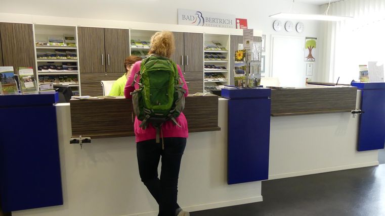 Eine Person mit einem grünen Rucksack steht an einem Empfangstresen. Der Bereich wirkt hell und modern, mit Regalen im Hintergrund.