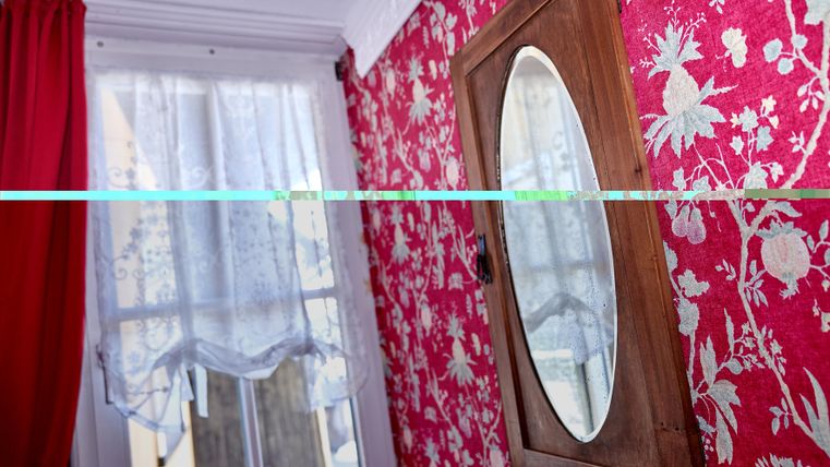 Ein Raum mit rot gemusterten Wänden und einem Holzspiegel. Vor dem Fenster hängt ein weißer Vorhang.