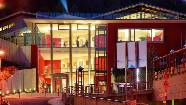 Ein modernes Gebäude mit großen Fenstern und roter Fassade. Es ist abends beleuchtet und umgeben von einer sanften Landschaft.