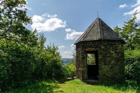 Ein steinernes Pavillon mit spitzem Dach steht in einer grünen Landschaft unter blauem Himmel.