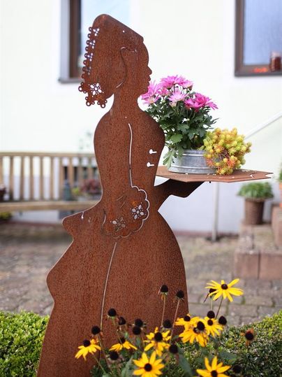 Eine dekorative Metallfigur, die eine Frau mit Blumen präsentiert. Im Hintergrund sind bunte Blumen und eine Holzbank zu sehen.