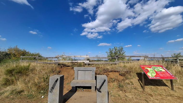 Eine Aussicht auf das Landschaftsschutzgebiet mit einer Betonbank und einem Informationsschild. Der Himmel ist klar mit einigen Wolken, und die Umgebung ist grün und trocken.