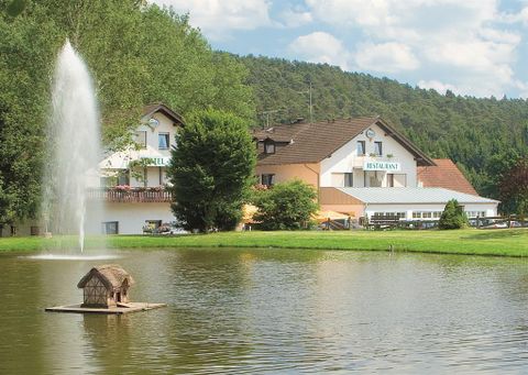 Ein ruhiger See mit einem kleinen Wasserbrunnen und einer gemütlichen Unterkunft im Hintergrund. Umgeben von Bäumen und Gras, bietet die Szene eine friedliche Atmosphäre.