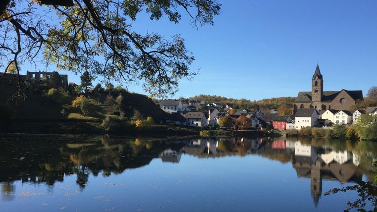 Ein malerisches Dorf am Fluss mit klarer Spiegelung im Wasser. Im Hintergrund sind Häuser und ein Kirchturm zu sehen, umgeben von Bäumen und blauem Himmel.