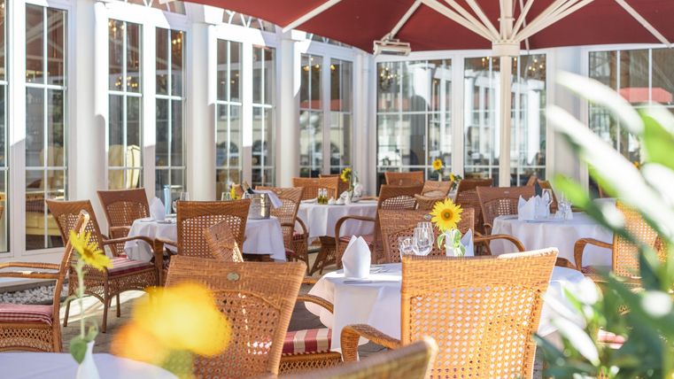 Ein gemütlicher Restaurantbereich mit schön gedeckten Tischen. Die Atmosphäre ist hell und einladend, umgeben von Pflanzen und Sonnenschirmen.