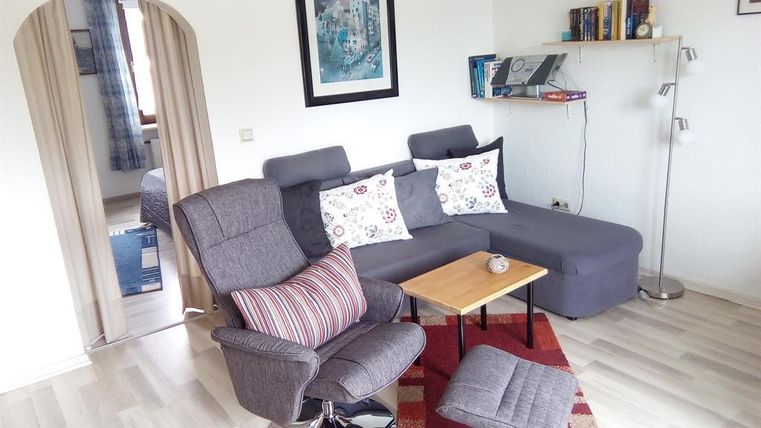 Ein gemütliches Wohnzimmer mit einem grauen Sofa und einem Sessel. Auf einem kleinen Tisch stehen eine Tasse und Dekoration, während Bücherregale im Hintergrund sichtbar sind.