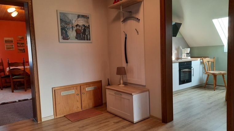 Ein heller Flur mit Holzfußboden und einem kleinen Sitzbereich. Im Hintergrund sieht man eine offene Küchenecke mit einer Kochstelle.