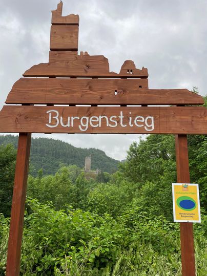 Ein hölzernes Schild mit der Aufschrift "Burgenstieg" vor einer grünen Landschaft. Im Hintergrund sind Hügel und eine Burgruine sichtbar.