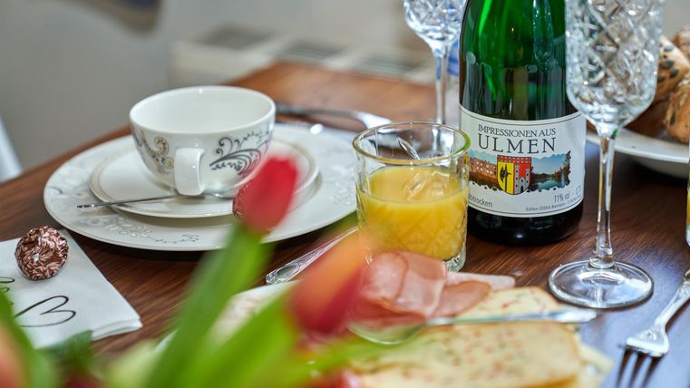 Ein liebevoll gedeckter Tisch mit einer Tasse, Orangensaft, Sekt und frischem Brot. Im Vordergrund sind Tulpen zu sehen.